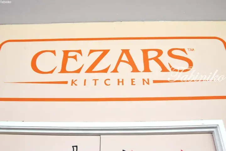 Close-up of the orange Ceazars Kitchen sign on a cream-colored wall, featuring the brand name in bold serif font.