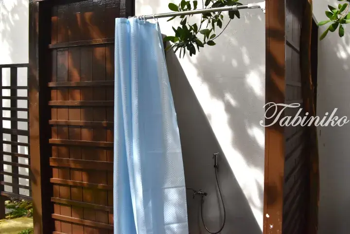 Outdoor shower area with a light blue curtain, wooden lattice privacy screen, and white wall at Tabiniko.