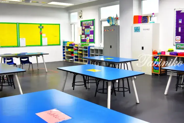 Bright classroom with blue tables, chairs, and colorful educational displays on the walls.
