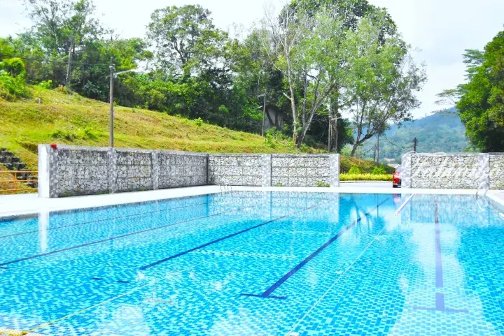 Outdoor swimming pool with blue tiles and lane ropes, surrounded by a gabion wall and lush green hillside.