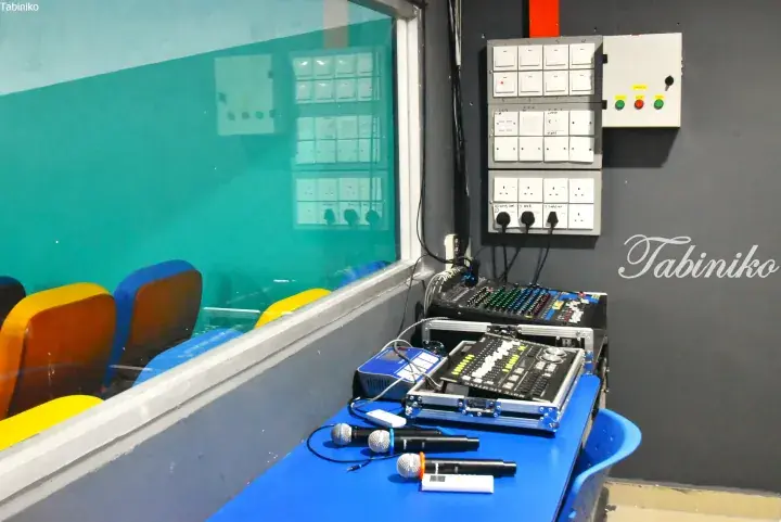 Audio mixing console and microphones on a blue desk in a control room overlooking a colorful hall.