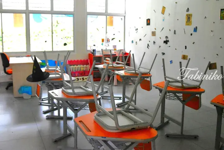 Empty classroom with orange desks and chairs stacked on top, decorated with children’s artwork on the walls.