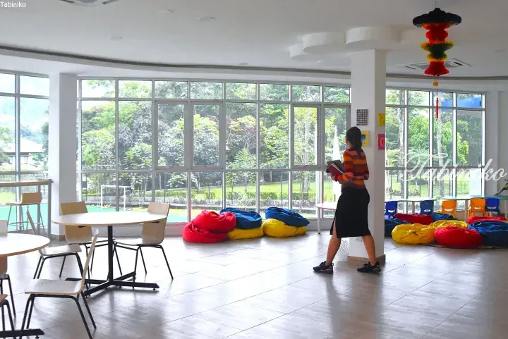 Bright classroom with large windows, colorful bean bags, and a teacher walking near a pillar.