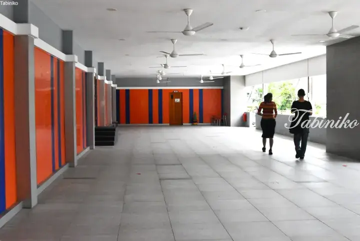 A spacious covered walkway with orange and blue striped walls, ceiling fans, and two people walking.