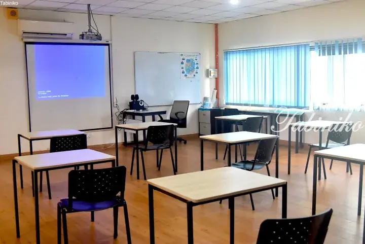 Bright classroom with wooden floors, white desks, black chairs, projector screen, and blue blinds.