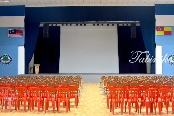 Tenby Schools auditorium with red chairs, blue curtains, Malaysian flag, and stage screen.