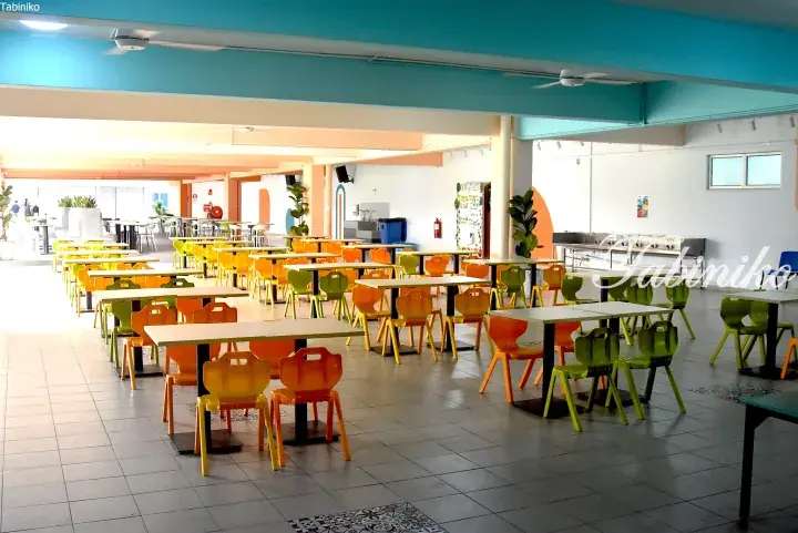 Bright school cafeteria with rows of small tables and colorful orange and green chairs under a blue ceiling.