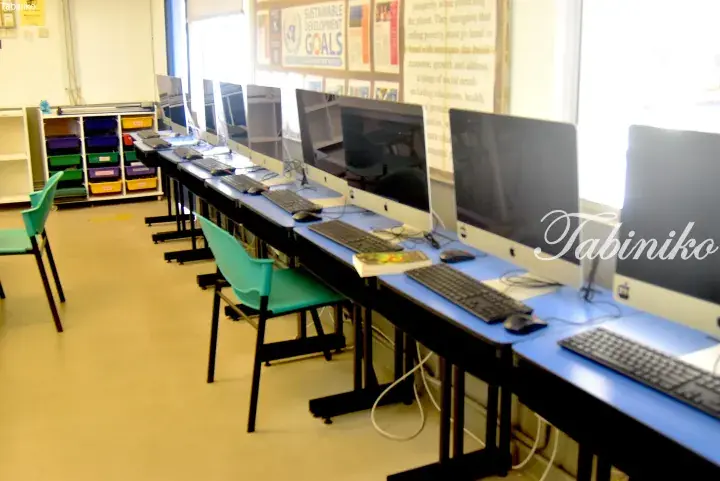 Row of Apple computers in a bright classroom with blue desks and green chairs near a Sustainable Development Goals poster.