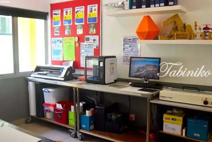 A modern workshop featuring a vinyl cutter, 3D printer, and computer setup with instructional posters on a red wall.