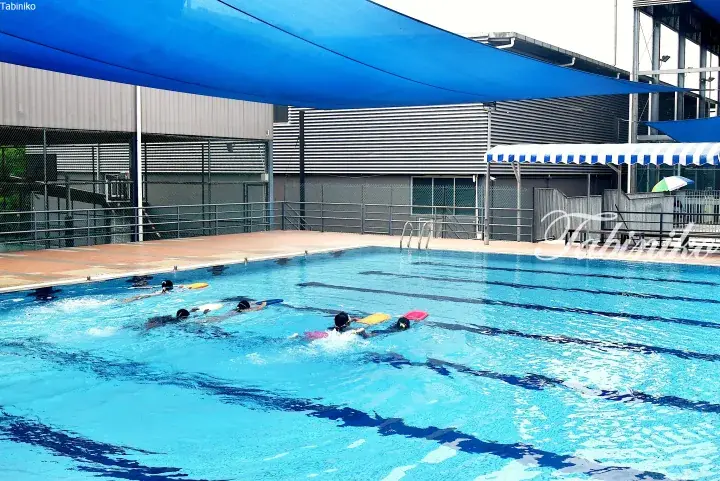 Children practicing swimming with kickboards in an outdoor pool under a blue shade canopy.