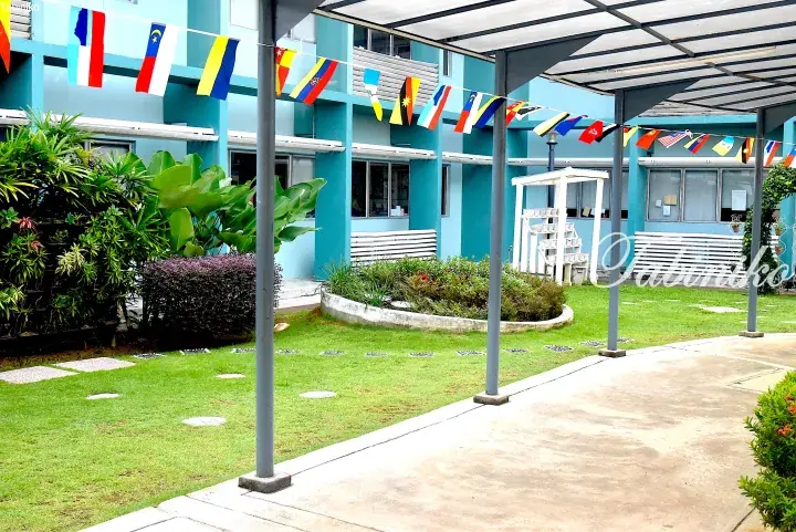Colorful international flags hang over a courtyard with a blue building, green lawn, and garden beds.
