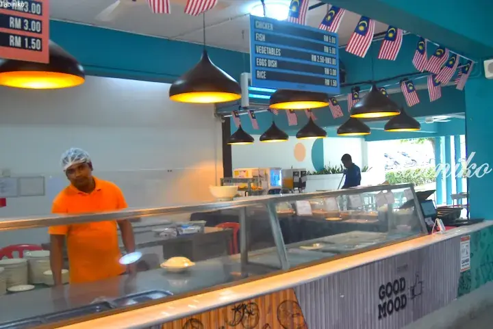 Staff in orange shirt serves customers at a modern Malaysian food stall with menu board and flags.