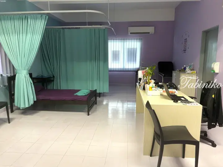 A clean clinic room with a patient bed behind green curtains, a desk with a laptop, and purple walls.