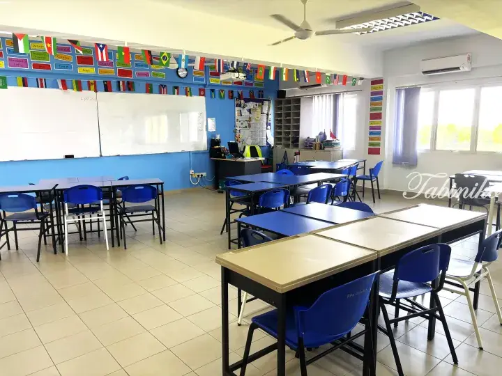 Bright classroom with blue chairs, beige desks, and international flags hanging above whiteboards.