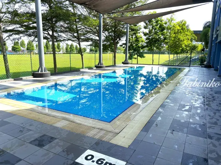 Outdoor swimming pool with blue tiles and shade canopy next to a green football field at a Malaysian property.