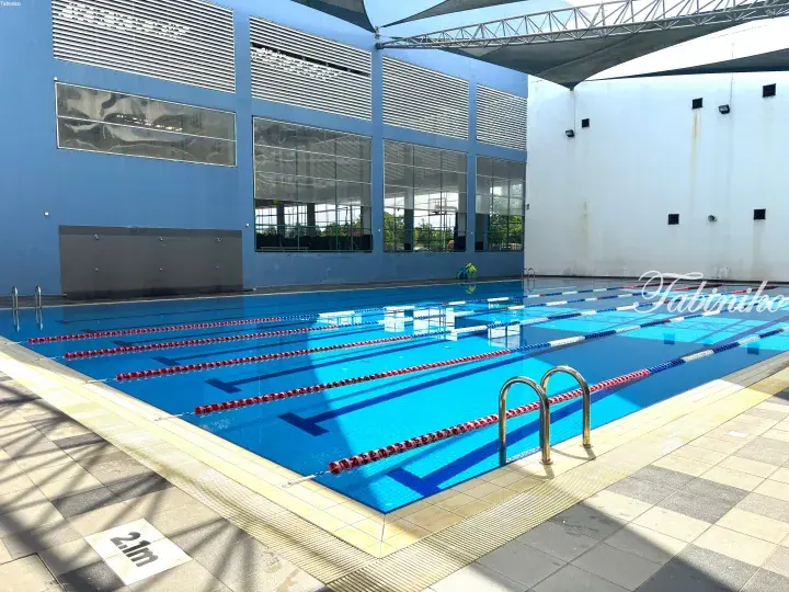 Modern indoor swimming pool with blue tiles, lane dividers, and large windows overlooking a sports court.