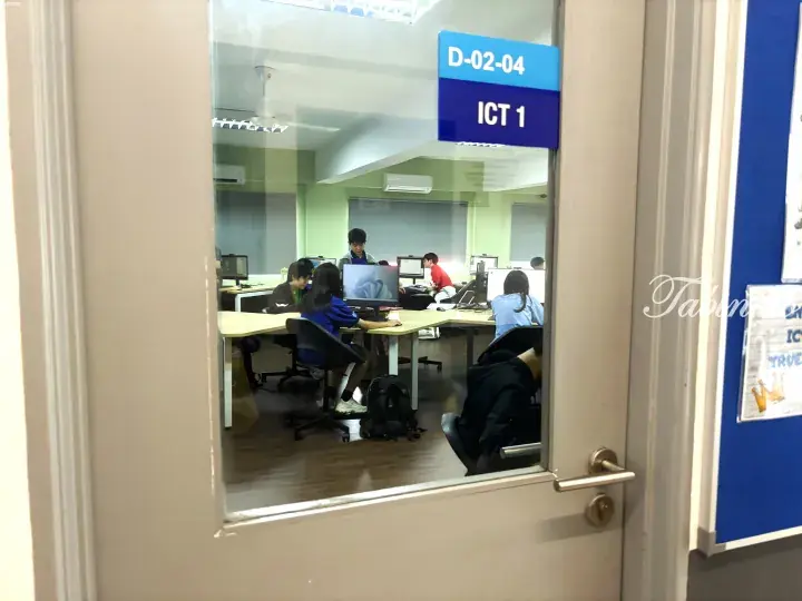 Students working on computers in a classroom labeled ICT 1, viewed through a glass door.