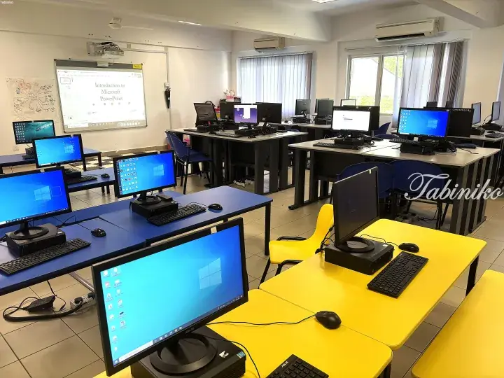 Modern computer lab with rows of desktops on blue and yellow tables, projector screen showing PowerPoint intro.