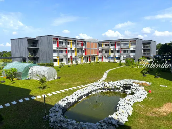 Modern grey dormitory building with colorful accents overlooking a landscaped garden with a pond and greenhouse.