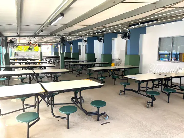 Spacious school cafeteria with rows of green tables and stools under a high ceiling with fans and string lights.