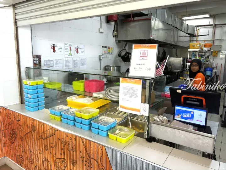 Cezars Kitchen food counter with colorful meal boxes, digital payment screen, and staff in orange uniform.