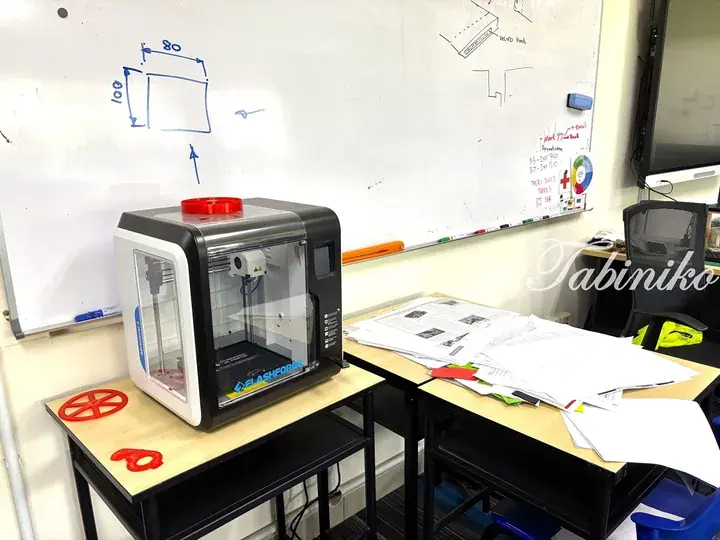FlashForge 3D printer on a desk with printed red objects and a whiteboard with diagrams in a classroom.