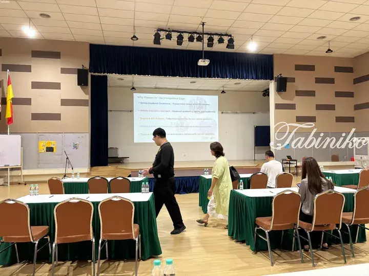 Attendees at an education seminar in a hall with a presentation screen showing school benefits.