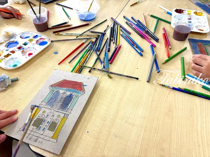 A child’s watercolor painting of a traditional building with a red roof on a wooden table surrounded by art supplies.