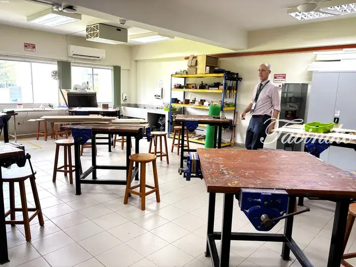 A man stands in a well-lit workshop with wooden workbenches, stools, and storage shelves.