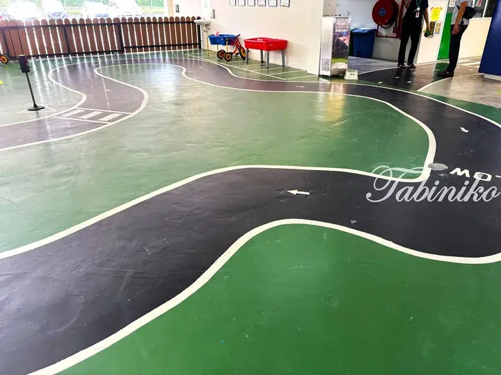 Indoor play area with a winding black road painted on green flooring, featuring a ‘Tabiniko’ logo and tricycles.