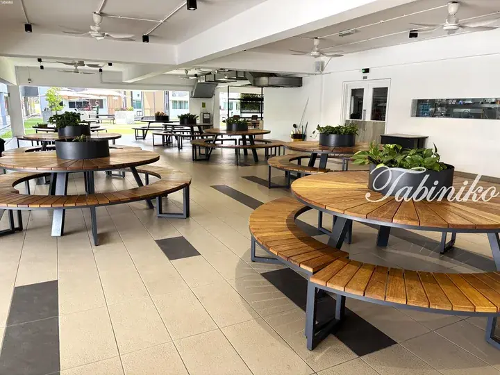 Modern communal dining area with circular wooden tables and curved benches in a bright, open-air space.