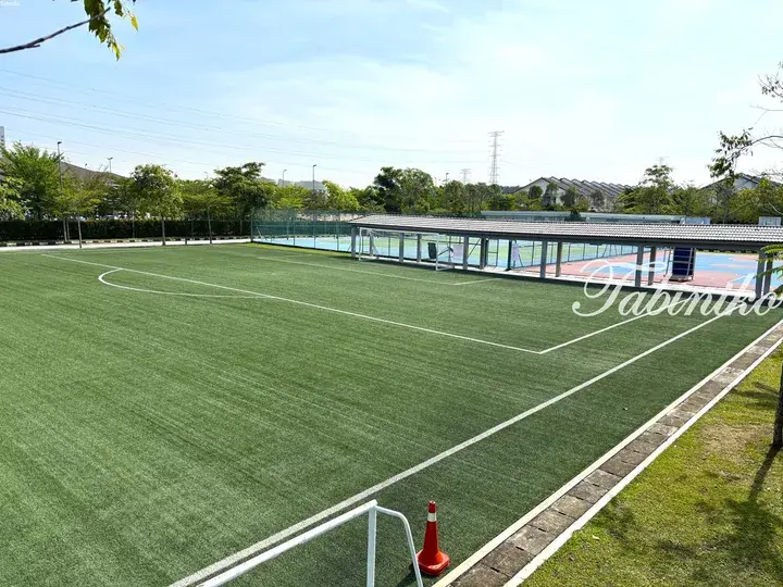 Green artificial turf soccer field with white boundary lines, adjacent to a covered basketball court in a Malaysian residential area.