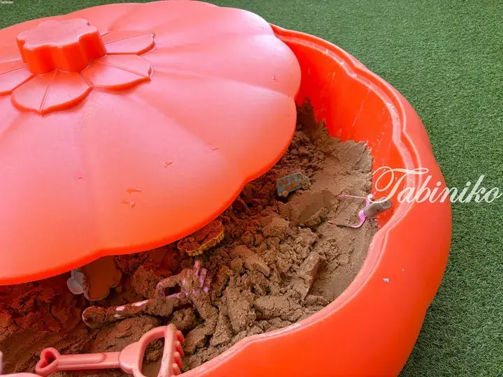 Close-up of a child’s orange pumpkin-shaped sandbox filled with sand and colorful plastic play tools on green artificial turf.