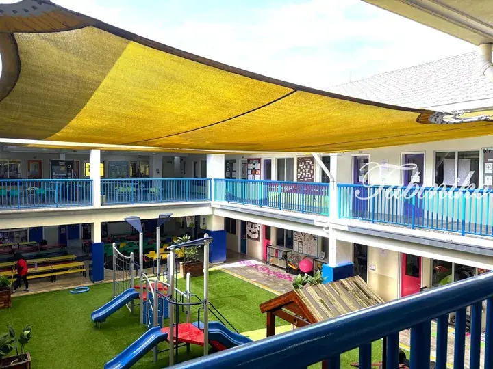 A school courtyard with a yellow shade sail, blue railings, and playground equipment on artificial grass.