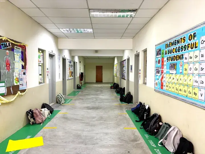 Empty school hallway with backpacks lined up on green floor mats and educational bulletin boards on the walls.