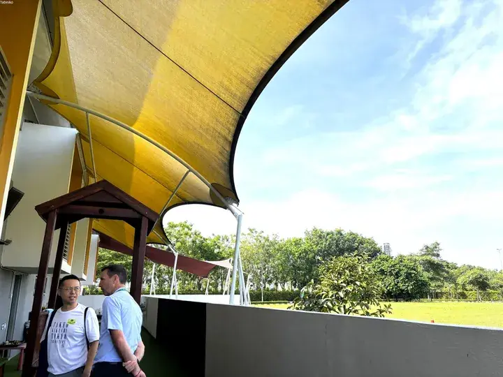 Two men standing under a large yellow shade sail at a property with a green lawn and trees.