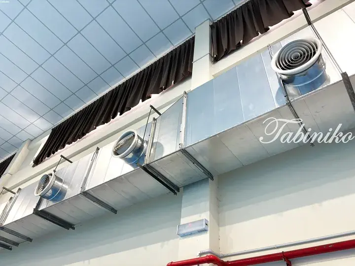 Galvanized ventilation ducts with circular outlets installed along a high ceiling in a commercial space.