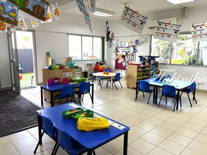 Bright classroom with blue tables and chairs, educational posters, and children’s artwork hanging from the ceiling.