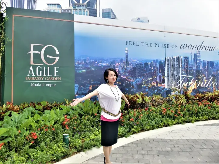 Woman posing in front of the Agile Embassy Garden Kuala Lumpur sign with lush garden and city skyline backdrop.