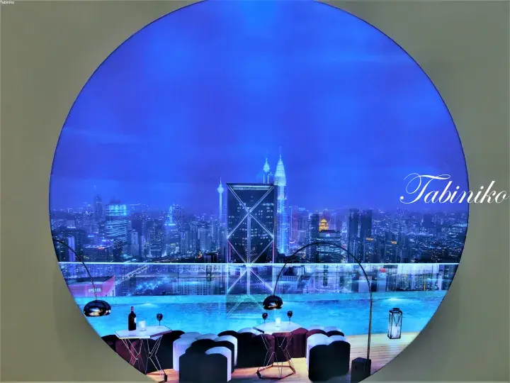 Night view of Kuala Lumpur skyline including Petronas Twin Towers seen through a circular window from a rooftop infinity pool.