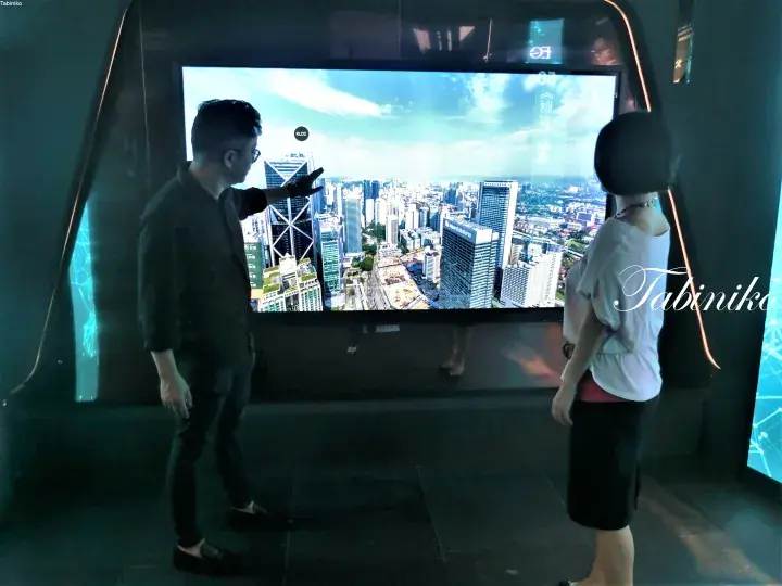 Couple viewing a large interactive display of Kuala Lumpur’s skyline, pointing at the KLCC landmark.
