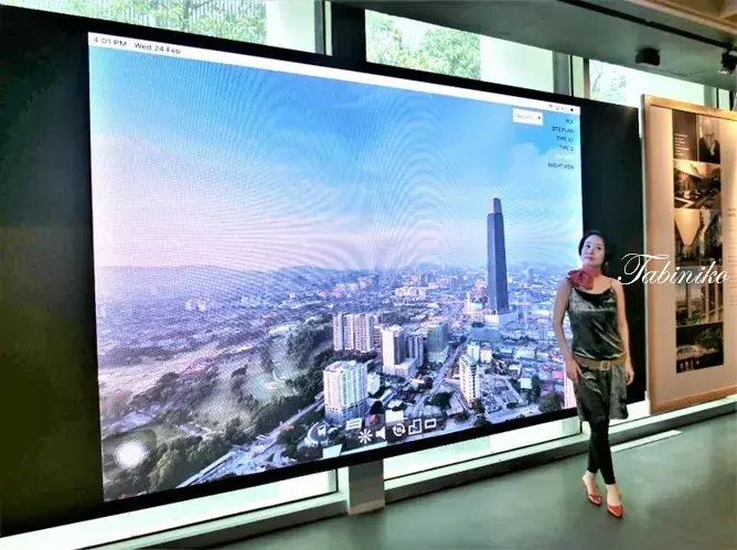 A woman stands beside a large digital display showing a panoramic view of Kuala Lumpur&rsquo;s city skyline.