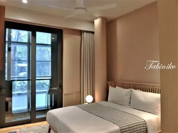 Modern hotel bedroom with a double bed, beige walls, and glass doors opening to a balcony.