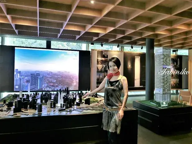 Woman posing at a property sales gallery with a large architectural model and cityscape display screen.