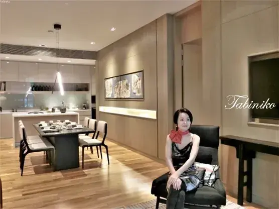 Woman in black dress and red scarf sitting in modern dining room with wooden floors and kitchen island.