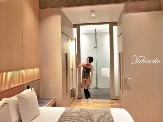 Woman in a modern hotel room opening a closet door near a glass-enclosed bathroom.