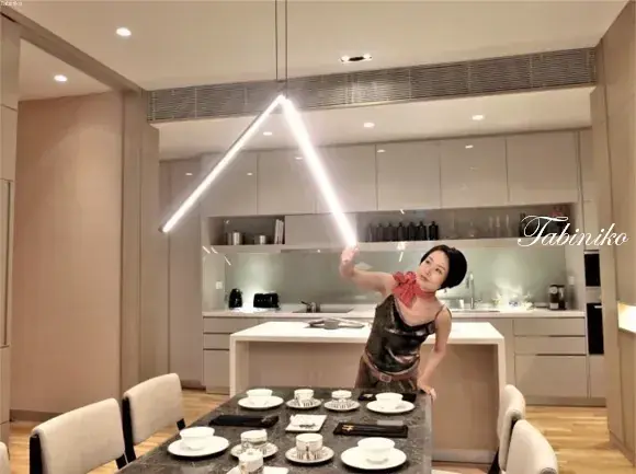 Woman posing playfully with a glowing triangular pendant light in a modern kitchen and dining area.