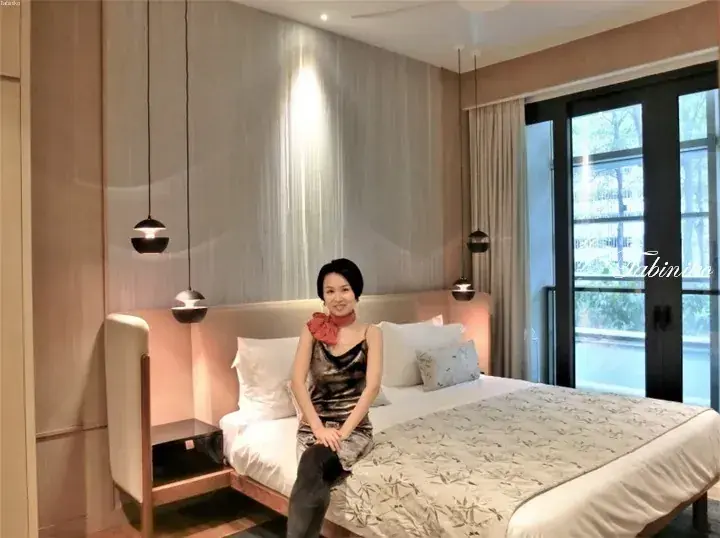Woman sitting on a modern hotel bed with wooden headboard and pendant lights in a well-lit room with glass doors.