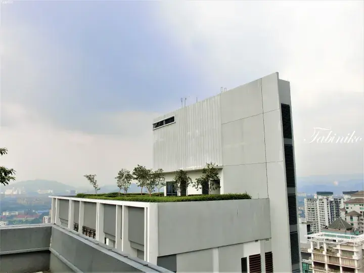 Modern grey building with rooftop garden and trees overlooking Kuala Lumpur cityscape and mountains.