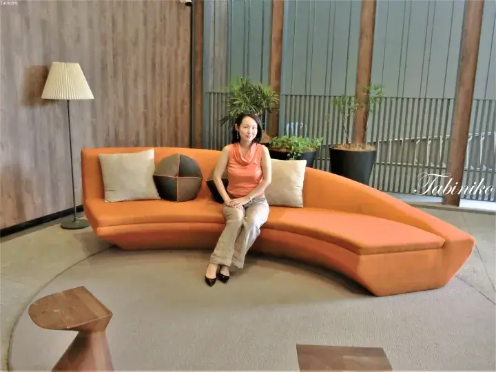 Woman sitting on a curved orange sofa in a modern hotel lobby with wooden accents and plants.